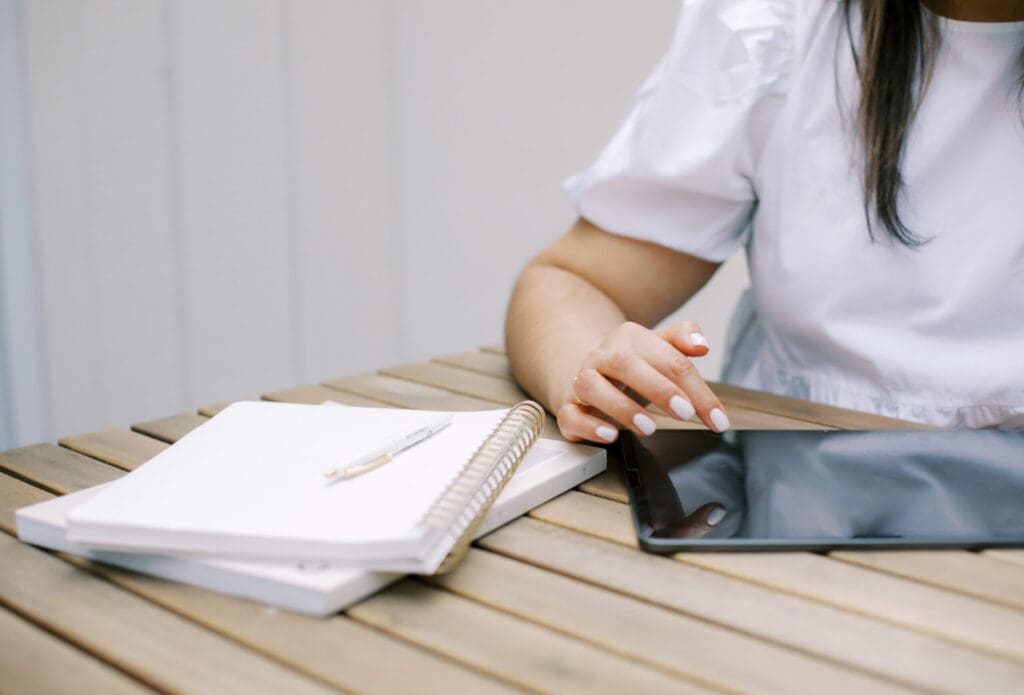 White woman working at outside wood table on Ipad with notebook next to her.
